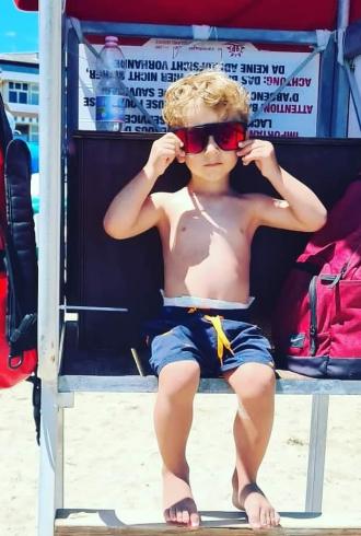 Kind am Strand mit Sonnenbrille auf einem Stuhl sitzend.